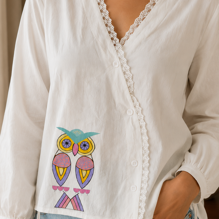 Woman wearing a white blouse with an owl design and lace details, standing indoors.